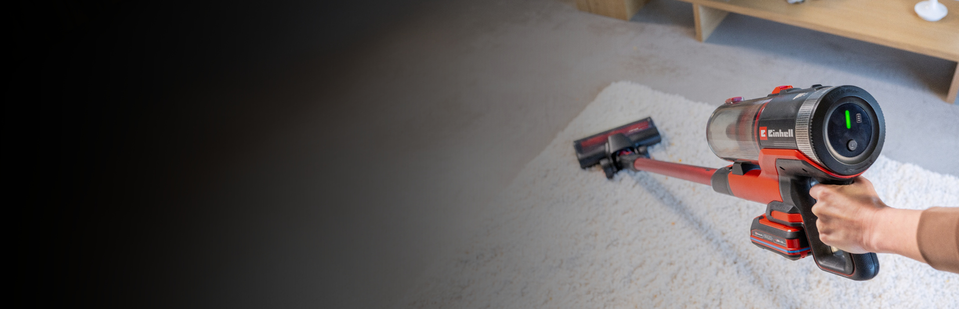 Das Bild zeigt einen kabellosen Staubsauger, der über einen hellen Teppich geführt wird. Der Staubsauger besitzt ein Display und auffällige rote Elemente, während im Hintergrund ein Teil eines Wohnzimmers mit Holzmöbeln erkennbar ist.