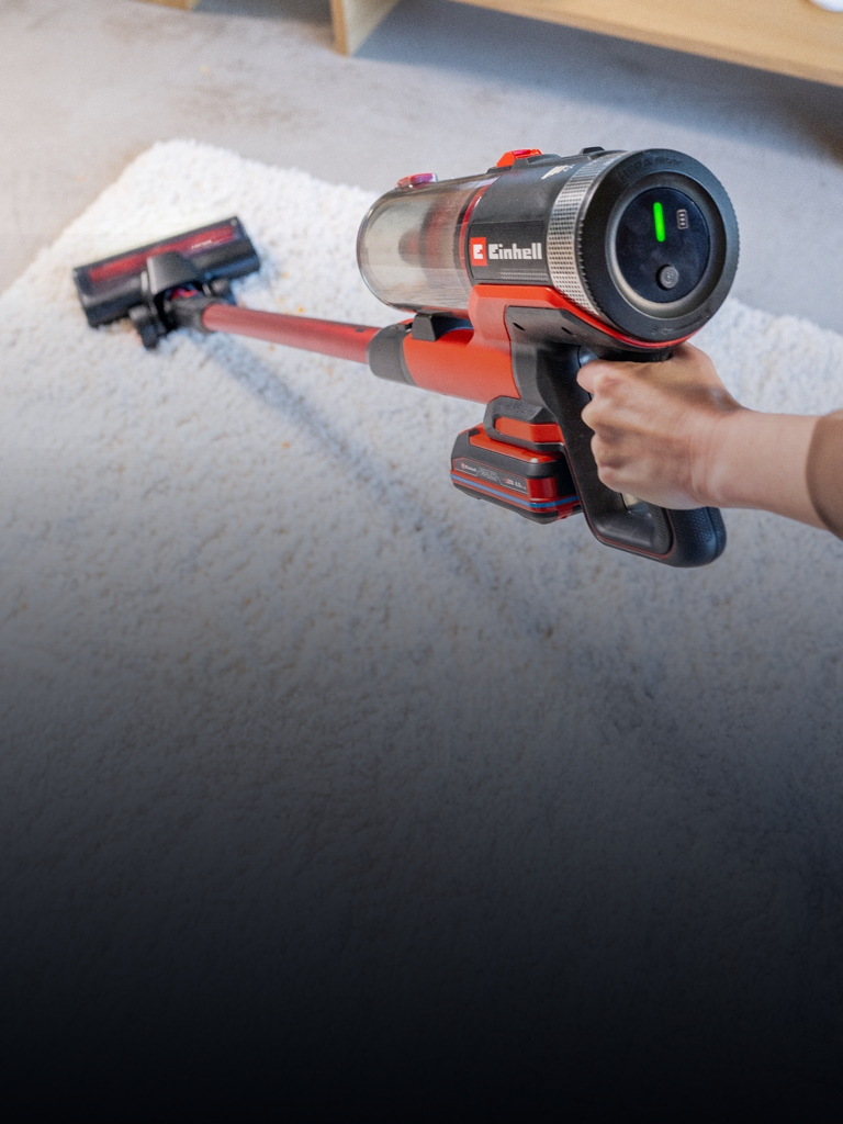 Das Bild zeigt einen kabellosen Staubsauger, der über einen hellen Teppich geführt wird. Der Staubsauger besitzt ein Display und auffällige rote Elemente, während im Hintergrund ein Teil eines Wohnzimmers mit Holzmöbeln erkennbar ist.