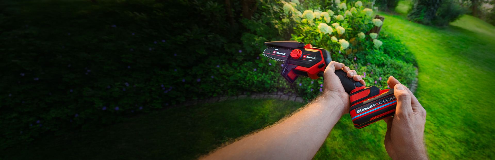 A person holding a red and black Einhell cordless pruning chainsaw in a green garden with blooming hydrangeas in the background.