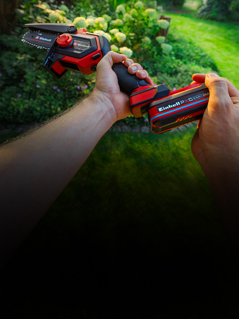 A person holding a red and black Einhell cordless pruning chainsaw in a green garden with blooming hydrangeas in the background.