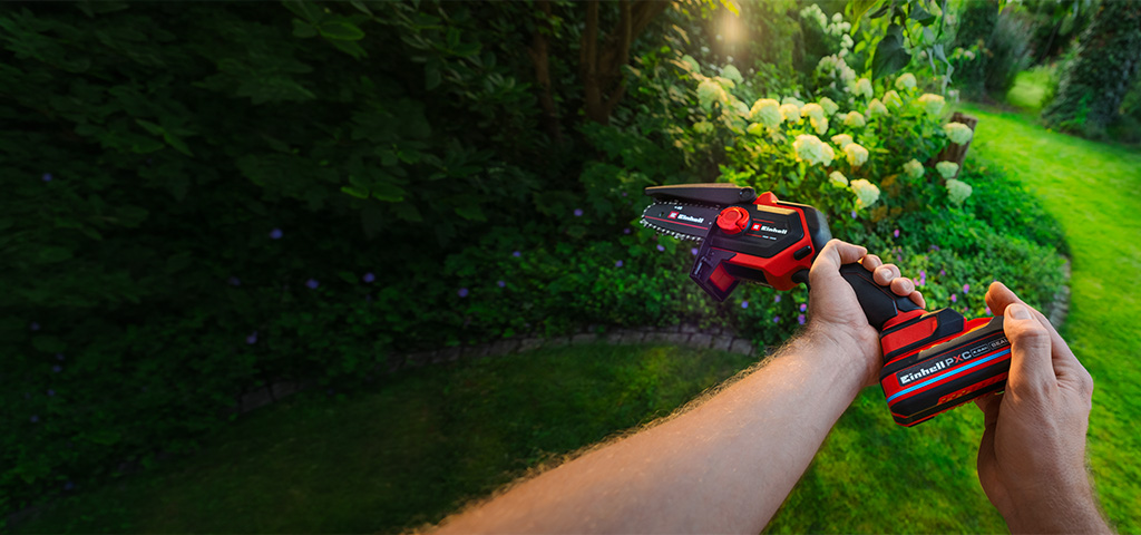 A person holding a red and black Einhell cordless pruning chainsaw in a green garden with blooming hydrangeas in the background.
