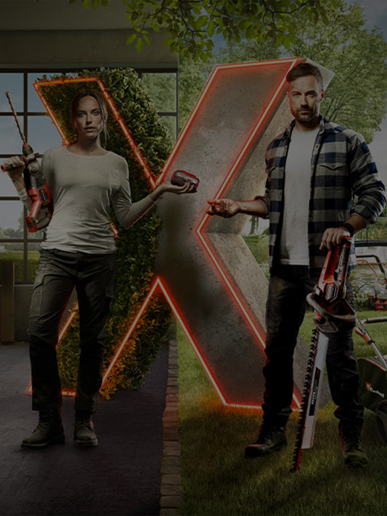 Two people stand in front of a large illuminated "X," surrounded by various cordless Einhell tools and garden equipment in a semi-open workshop and garden area.
