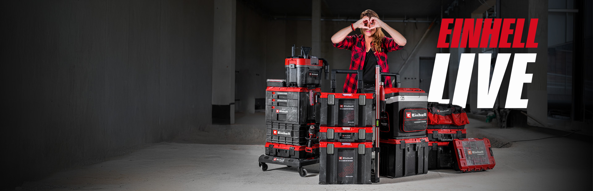 Person stands behind stacked Einhell tool cases on a construction site, making a heart with hands, next to the text “EINHELL LIVE”.