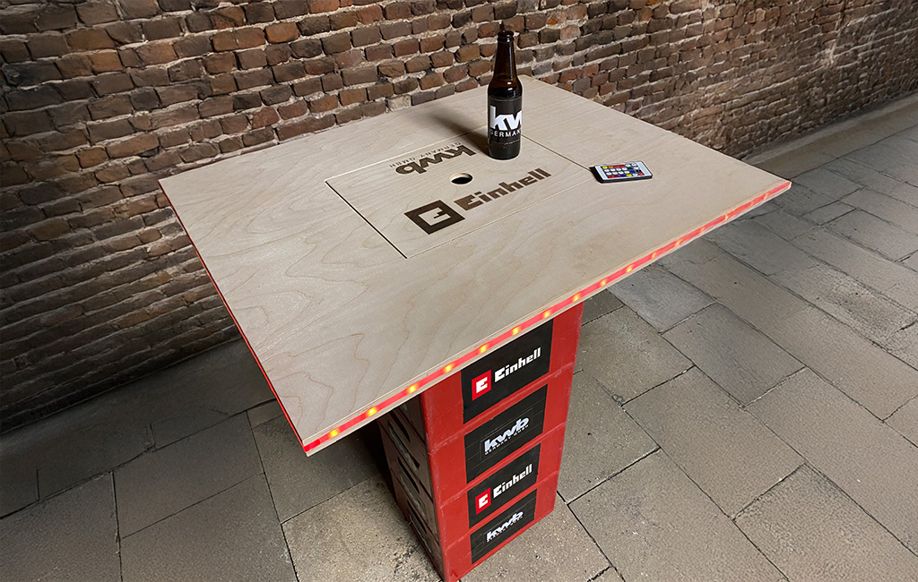 Ein selbstgebauter Stehtisch aus Holz mit LED-Leiste, auf einem Sockel mit Einhell- und kwb-Logos. Auf dem Tisch stehen eine Bierflasche und eine Fernbedienung. In die Tischplatte ist „Einhell“ und „kwb GERMANY GMBH“ gelasert.