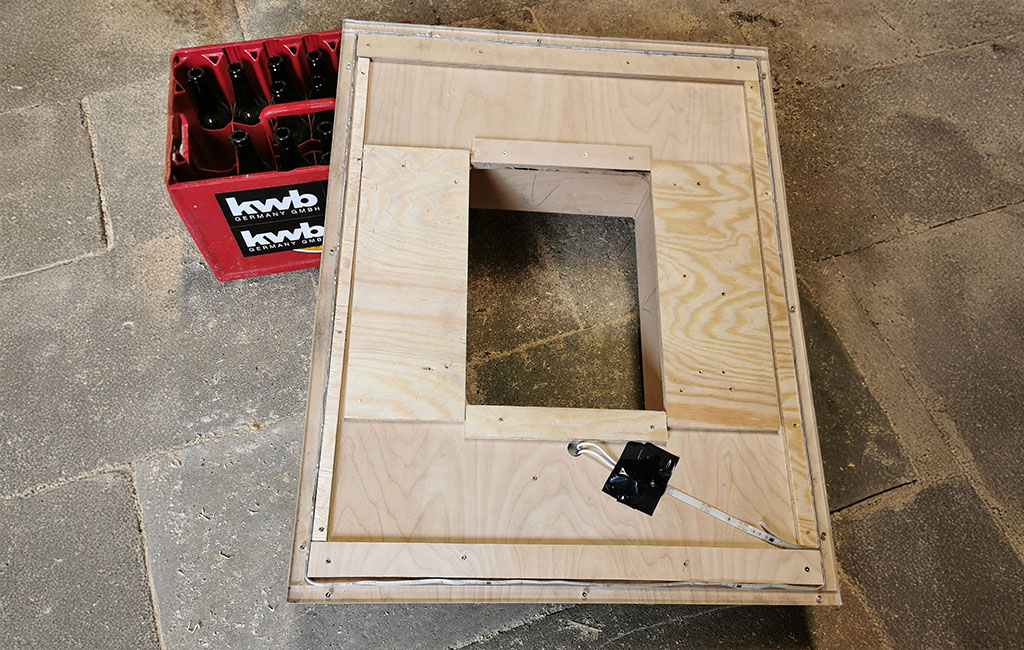 Unterseite einer Holzplatte mit quadratischem Loch, Kabel und LED-Streifen. Daneben steht eine leere kwb-Flaschenkiste.
