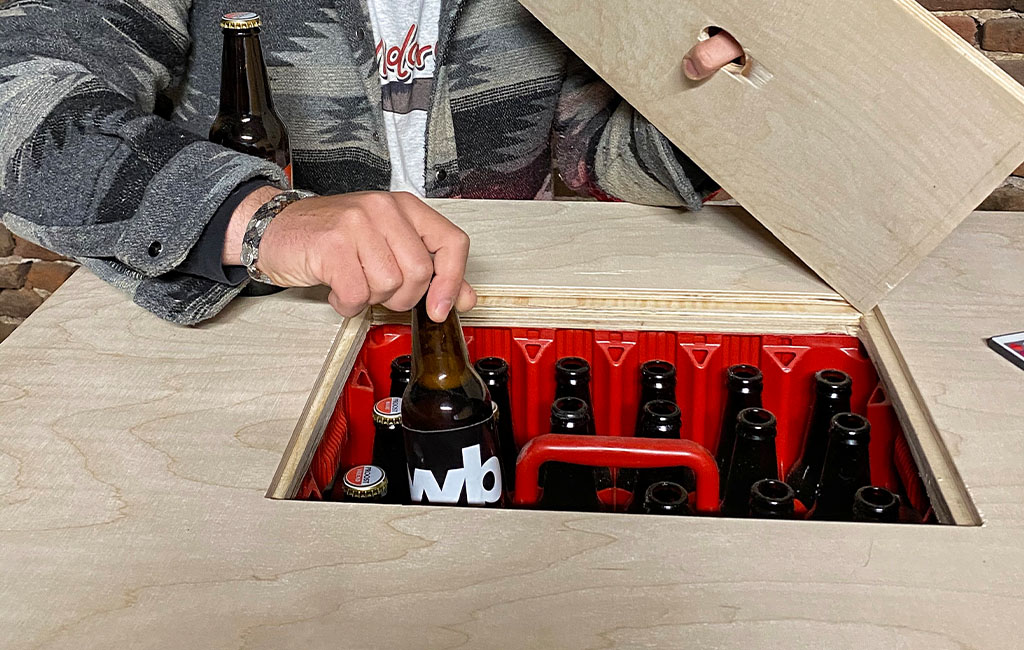 Eine Person hebt die mittlere Abdeckung des Tisches ab und greift nach einer Bierflasche in einer roten Getränkekiste im Inneren.