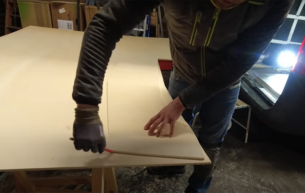 Mann zeichnet mit Bleistift und Holzleiste die geschwungene Form auf eine Sperrholzplatte.