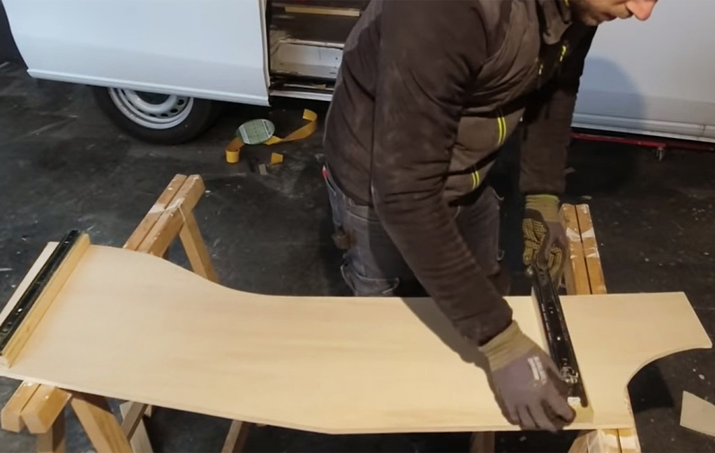 Mann montiert eine Metallschiene auf einem gebogenen Holzbrett vor einem Camper.