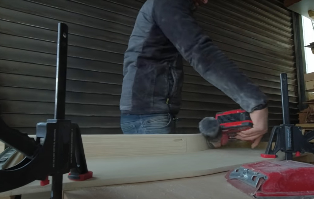 Mann schleift mit einem roten Akku-Exzenterschleifer eine Holzplatte, die auf einem Arbeitstisch eingespannt ist.