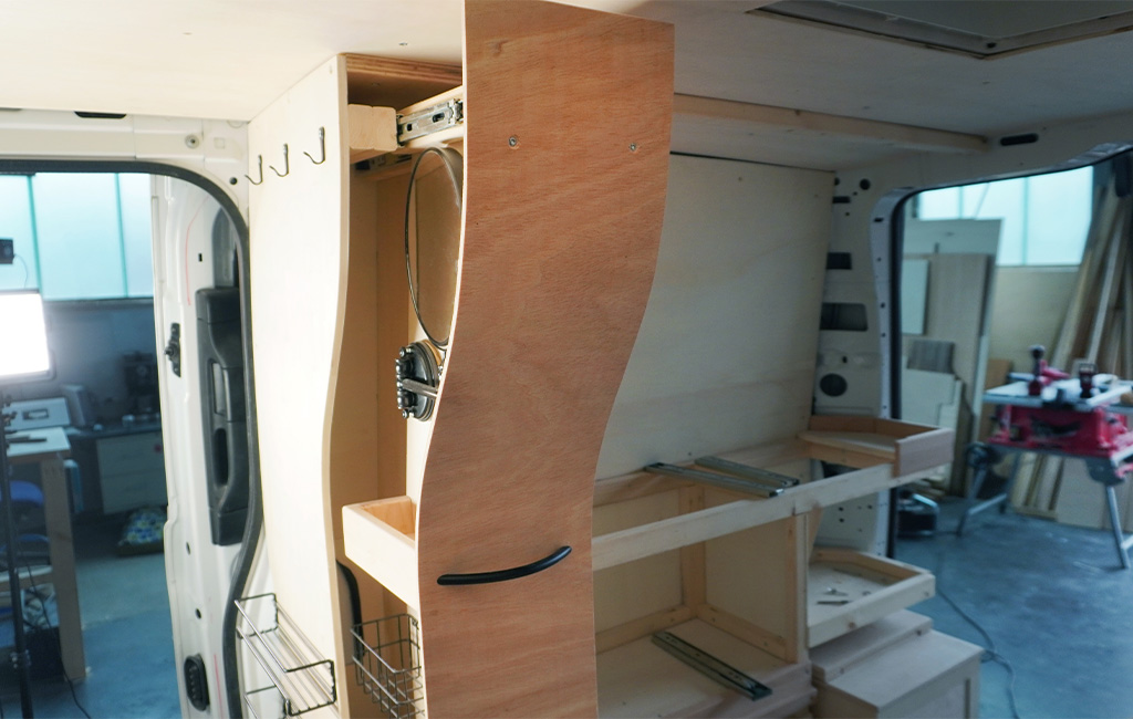 Eingebauter Badschrank im Camper mit geschwungener Front, Spiegel, Drahtkörben und Regalfächern.