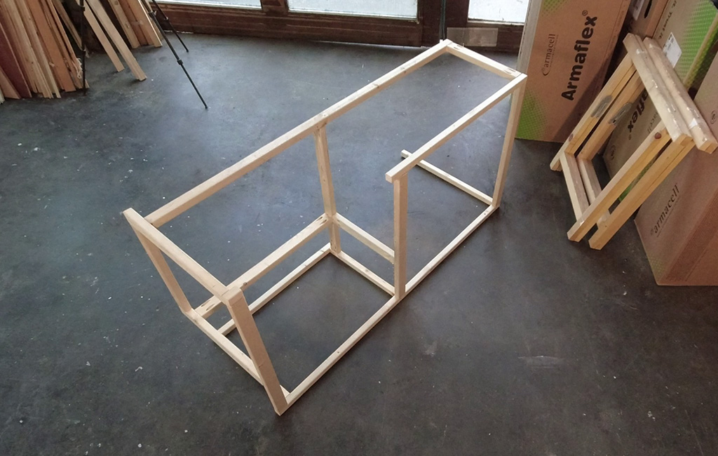 Leicht schräg aufgenommener Holzrahmen auf Werkstattboden, im Hintergrund Holzlatten und Kartons.