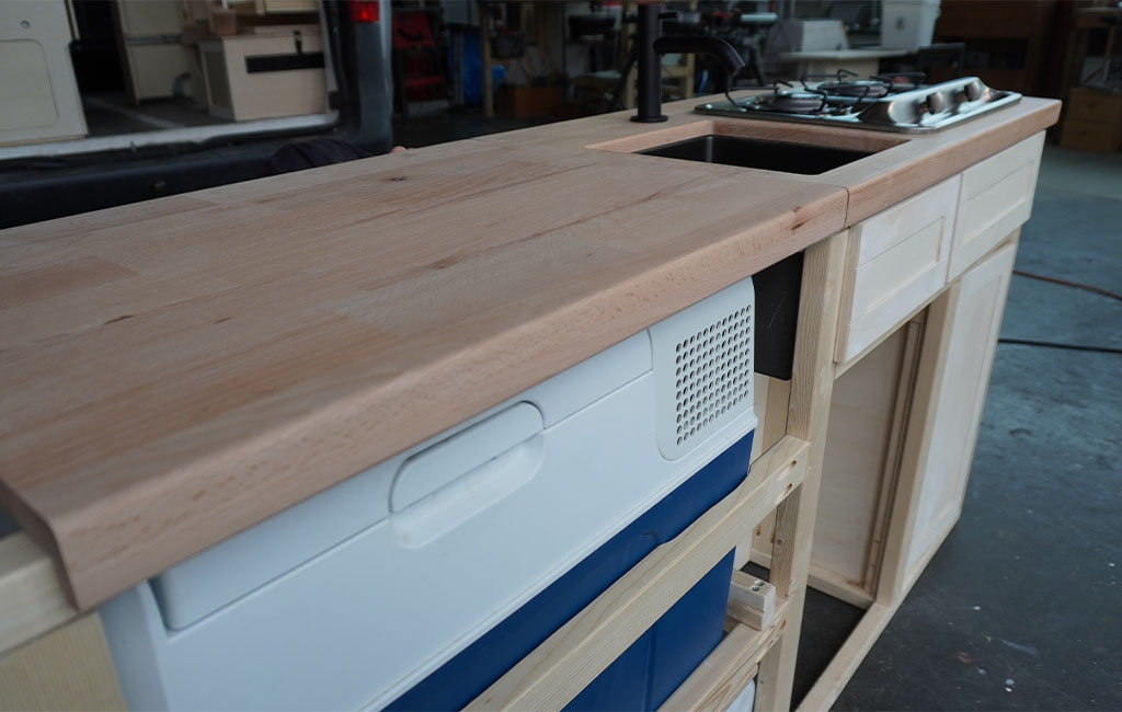 Küchenzeile im Bau mit Holzarbeitsplatte, darunter Kühlbox und offenes Regalsystem aus Holz.