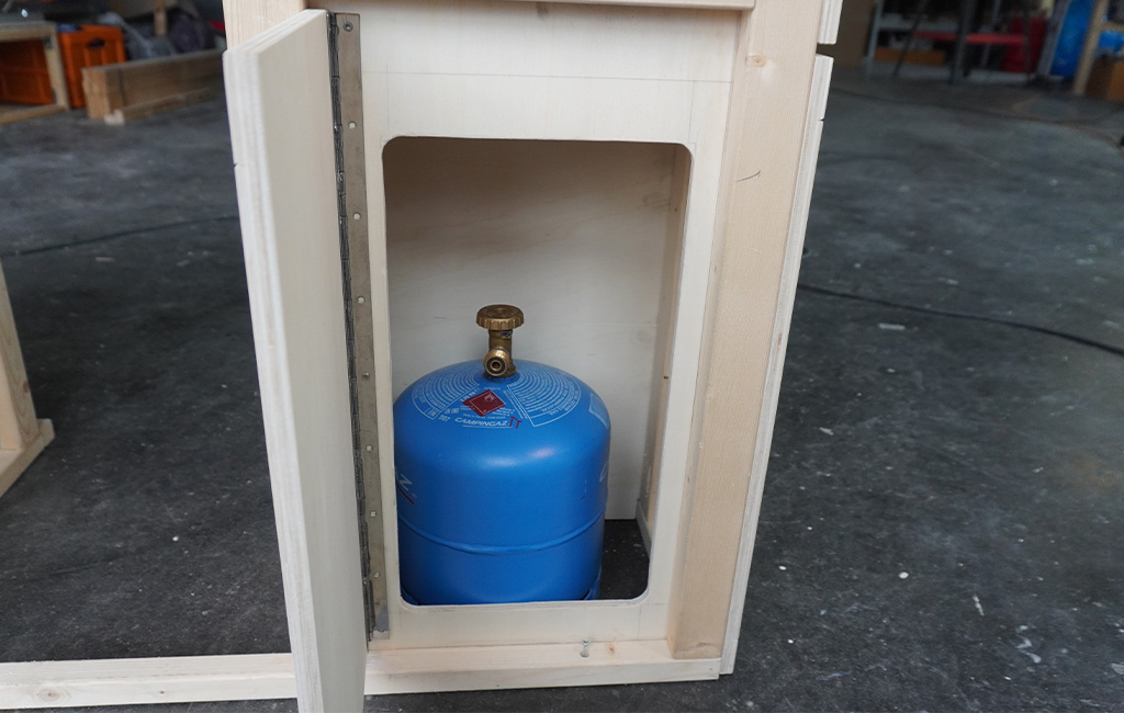 Eine blaue Gasflasche steht in einem offenen Holzfach eines Küchenmöbels.