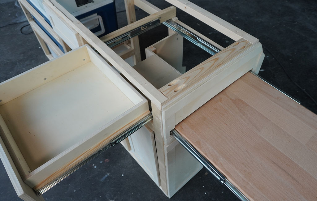 Zwei ausgezogene Schubladen mit Schienen in einer Küchenzeile im Aufbau, eine davon mit Holzplatte als Tisch.
