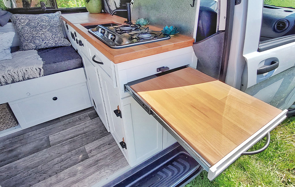 Fertig ausgebaute Camperküche mit Holzarbeitsplatte, Gasherd, Spüle und ausziehbarem Tisch bei geöffneter Seitentür.