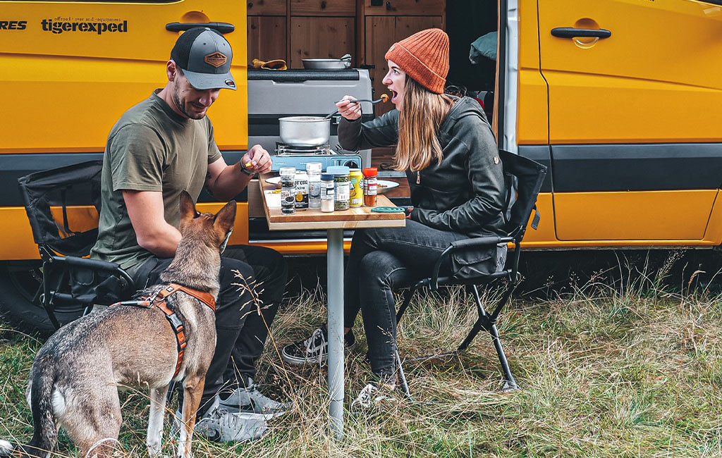 Ein Mann und eine Frau essen an einem Campingtisch vor einem gelben Van, ein Hund steht daneben.