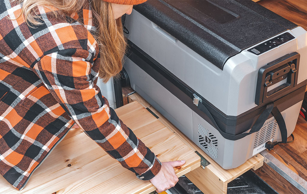 Eine Frau zieht eine ausklappbare Holzplatte neben einer großen Kühlbox im Camper heraus.