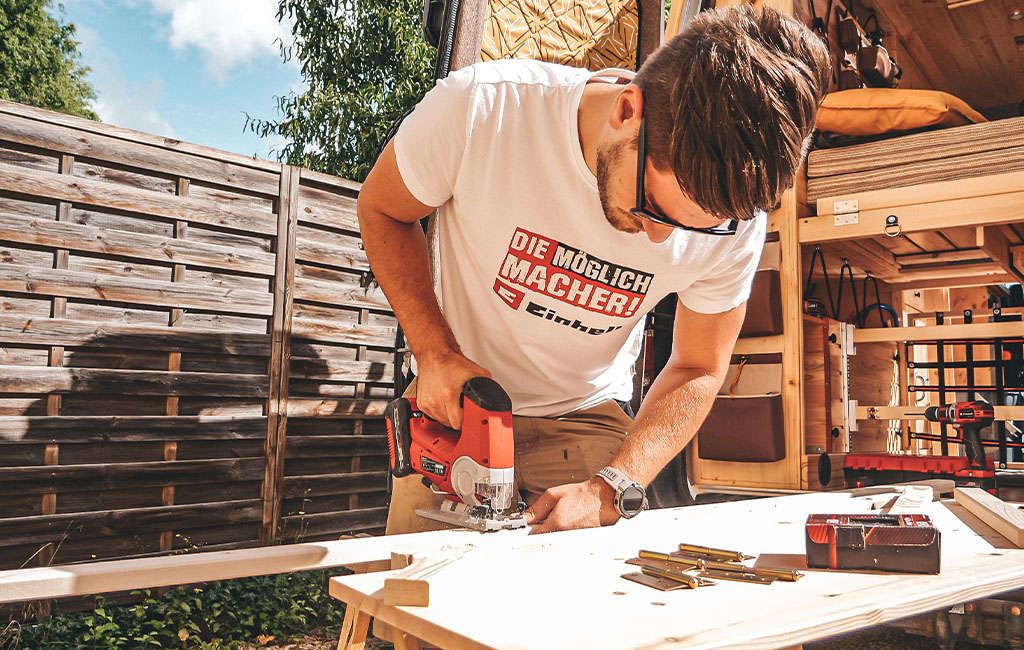 Ein Mann mit Einhell-T-Shirt sägt mit einer Stichsäge eine Holzplatte vor dem Camper.