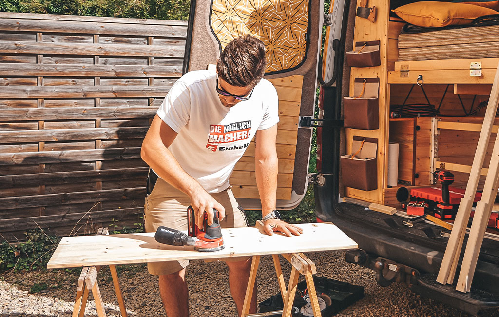 Ein Mann schleift mit einem Einhell Schleifer eine Holzplatte auf Böcken vor dem Camper.