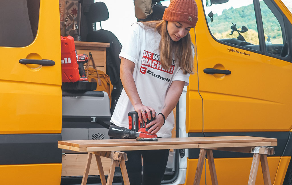 Eine Frau schleift draußen vor einem gelben Camper eine Holzplatte mit einem Einhell Schleifgerät.