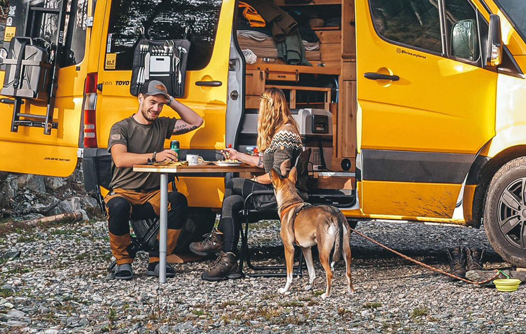 Ein Paar sitzt vor einem offenen Camper am Klapptisch, ein Hund steht davor.