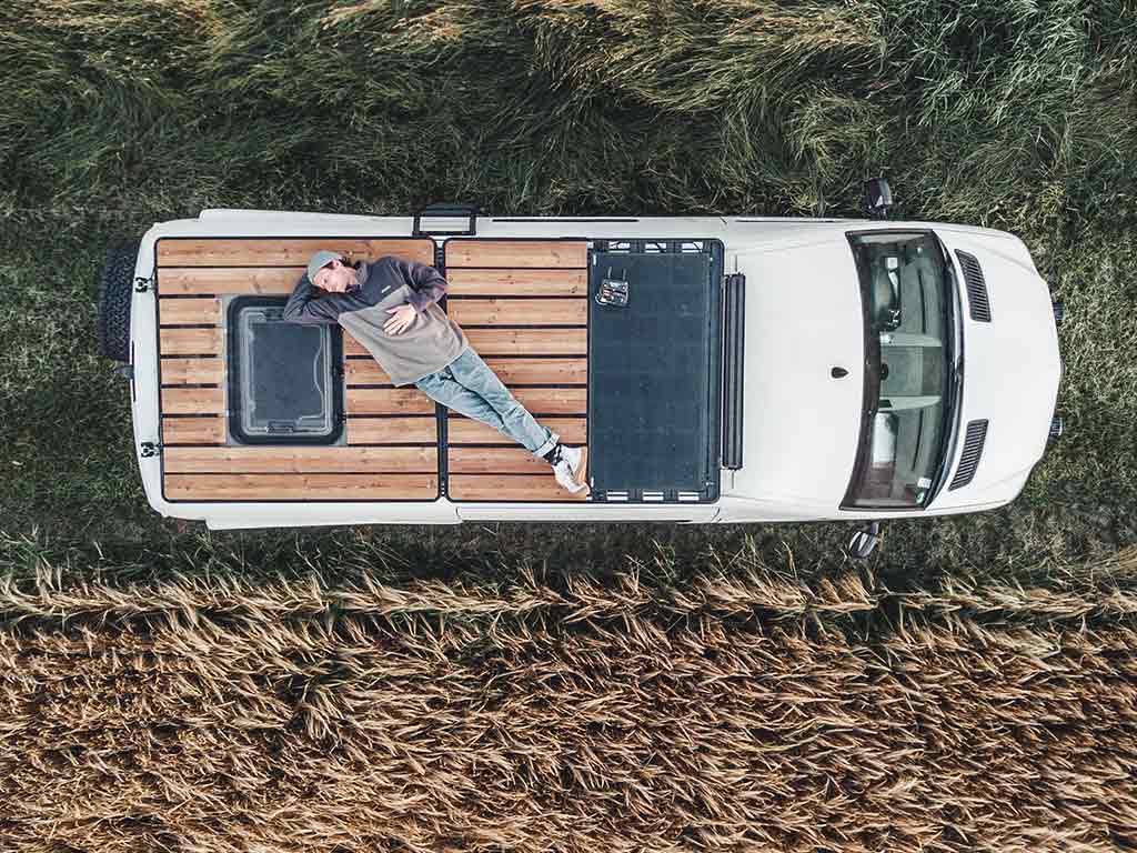 Person liegt entspannt auf einer Holzterrasse auf dem Dach eines Campers neben einem Getreidefeld.