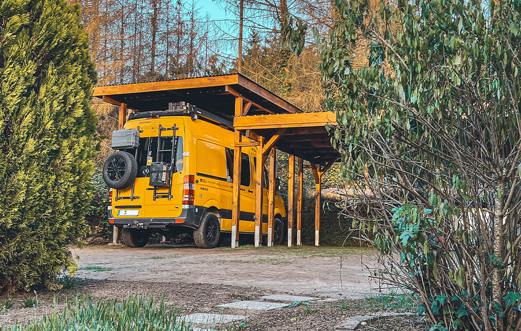 Ein gelber Van steht unter einem offenen Carport aus Holz, umgeben von Bäumen und Büschen.