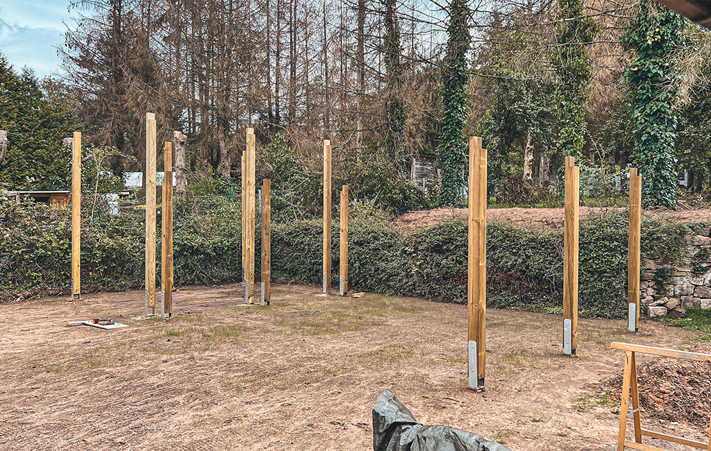 Mehrere senkrechte Holzpfosten mit Metallfüßen stehen auf einer ebenen Fläche, Baustelle im Hintergrund.