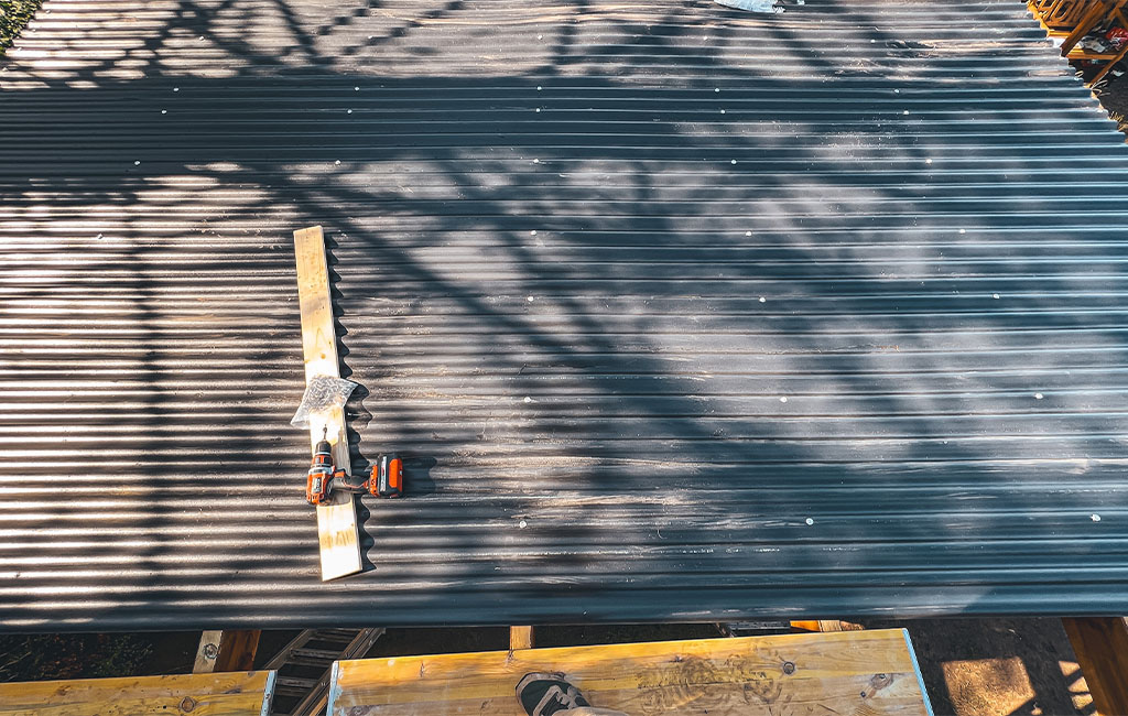 Blick von oben auf ein montiertes Carport-Wellblechdach mit Akku-Bohrschrauber, Holzlatte und Schrauben.