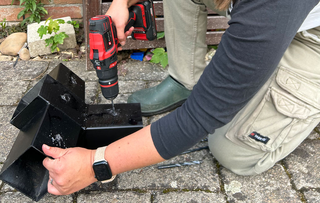 Eine Frau bohrt mit einem Einhell Akku-Bohrschrauber Löcher in ein schwarzes Metallverbindungsstück auf einem Pflasterboden.