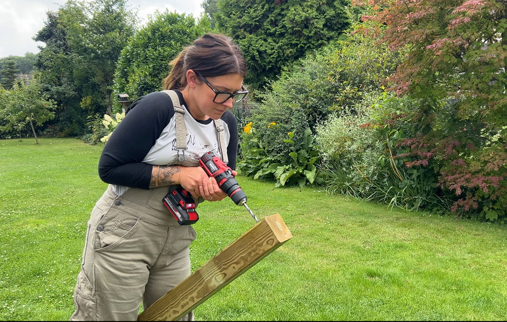 Eine Frau bohrt mit einem Einhell Akku-Bohrschrauber ein Loch in das Ende eines Holzbalkens im Garten.