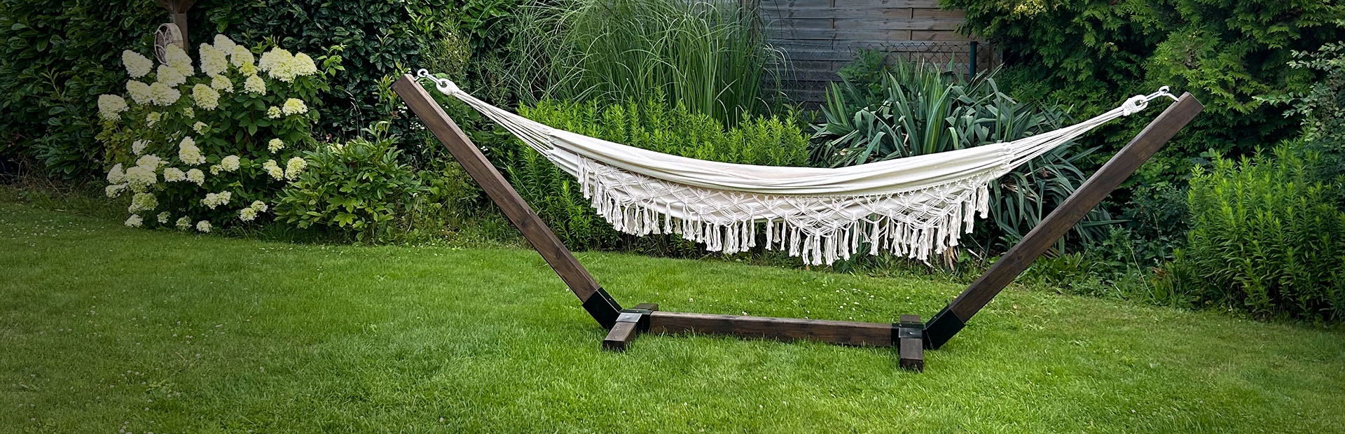 Eine cremefarbene Hängematte mit Fransen hängt in einem dunklen Holzgestell auf einer Rasenfläche im Garten.