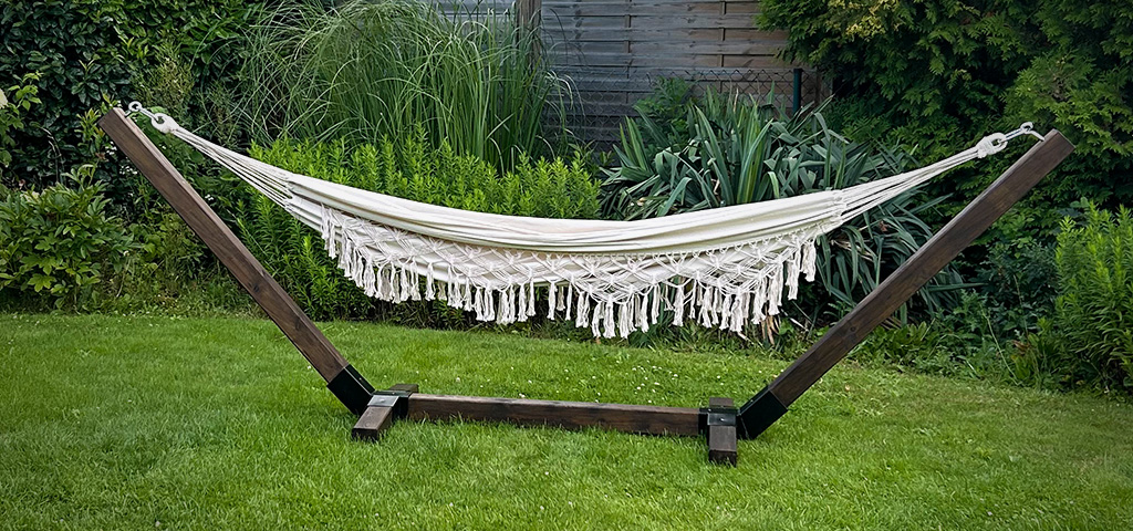 Eine cremefarbene Hängematte mit Fransen hängt in einem dunklen Holzgestell auf einer Rasenfläche im Garten.
