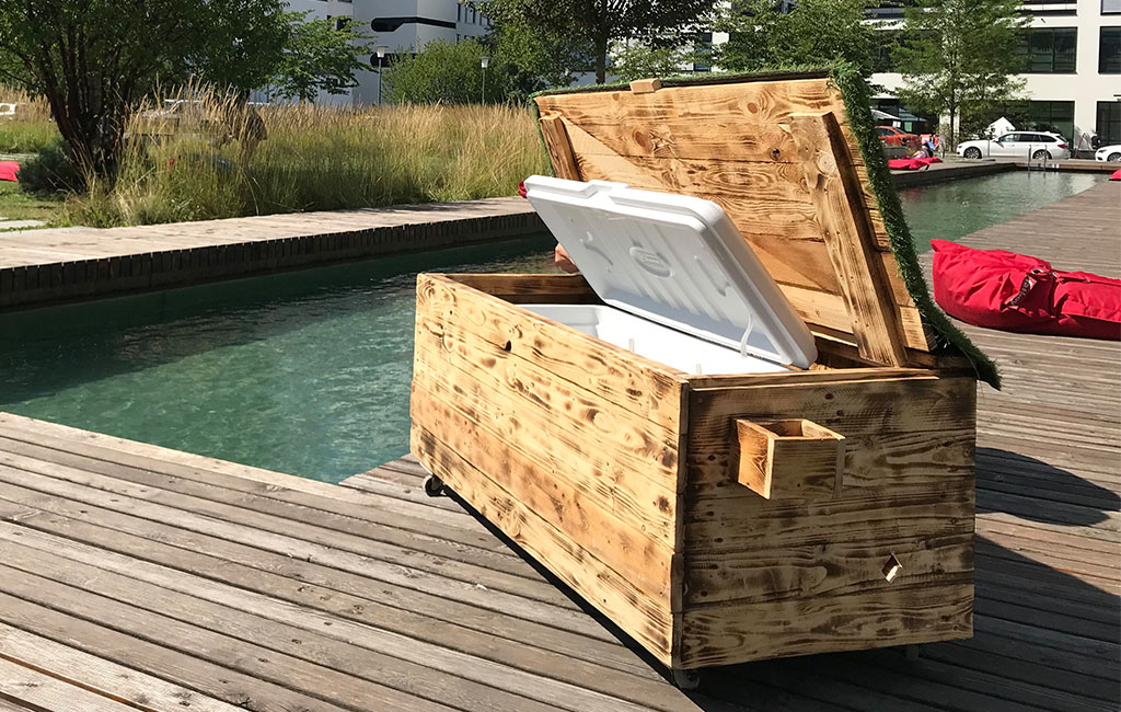 Eine große Holztruhe mit aufgeklapptem Deckel steht auf einer Holzterrasse neben einem Wasserbecken. Im Inneren befindet sich eine weiße Kühlbox.