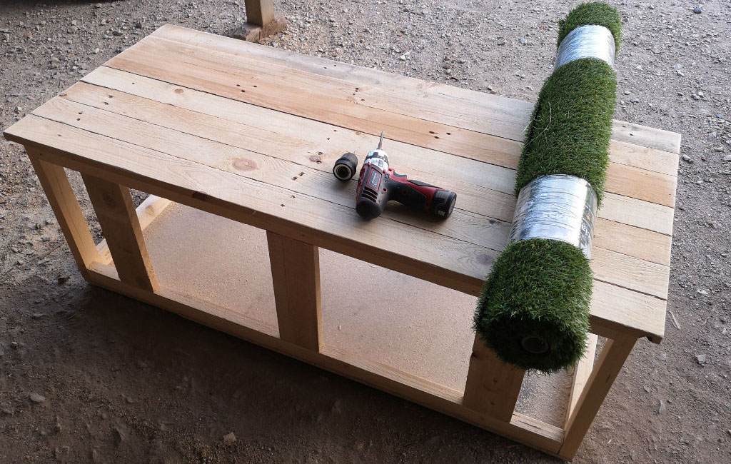 Ein einfacher Holztisch steht in einer Werkstatt. Auf dem Tisch liegen eine Bohrmaschine und eine Rolle Kunstrasen.