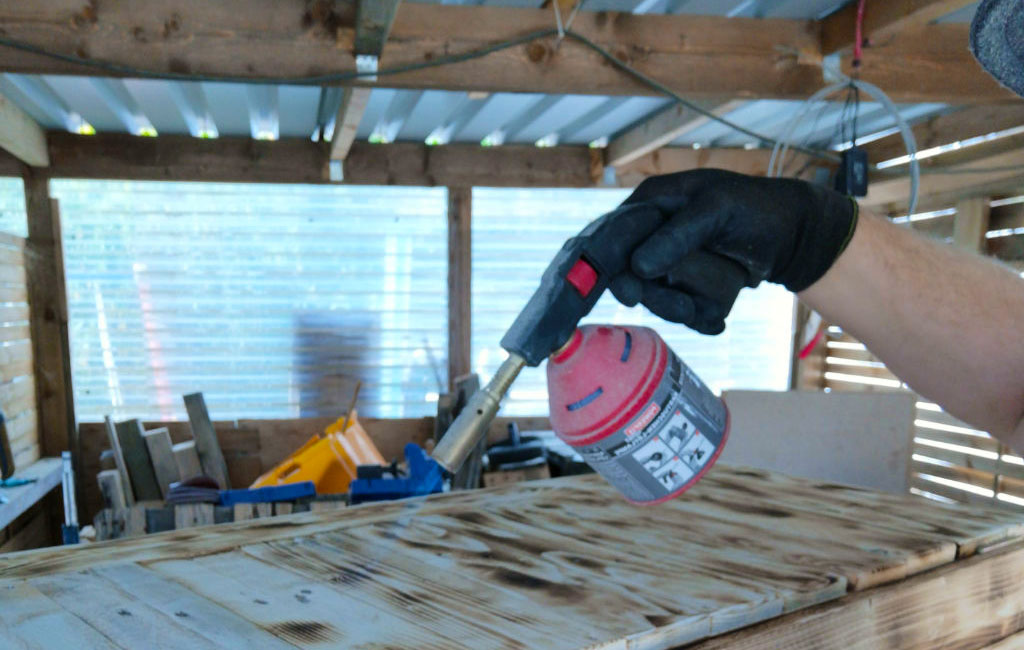 Eine Person hält eine Lötlampe mit roter Gaskartusche und brennt Holz in einer Werkstatt an.