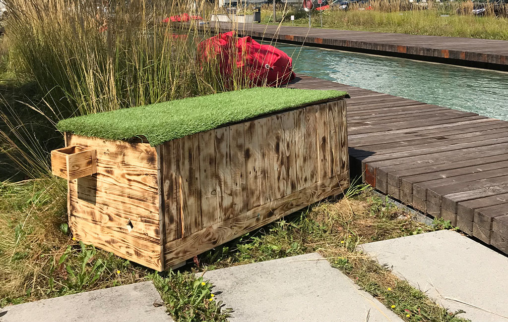 Die fertige Holztruhe mit Kunstrasen-Deckel steht auf einer Wiese neben einem Holzsteg.