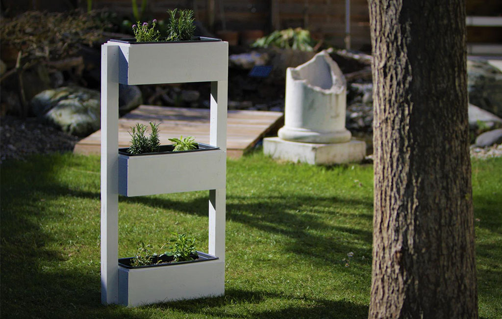 Ein weißer, rechteckiger Kräutergarten mit drei übereinander angeordneten Pflanzkästen im Garten. Im Hintergrund ein Baum, eine zerbrochene Steinsäule und ein Holzsteg.