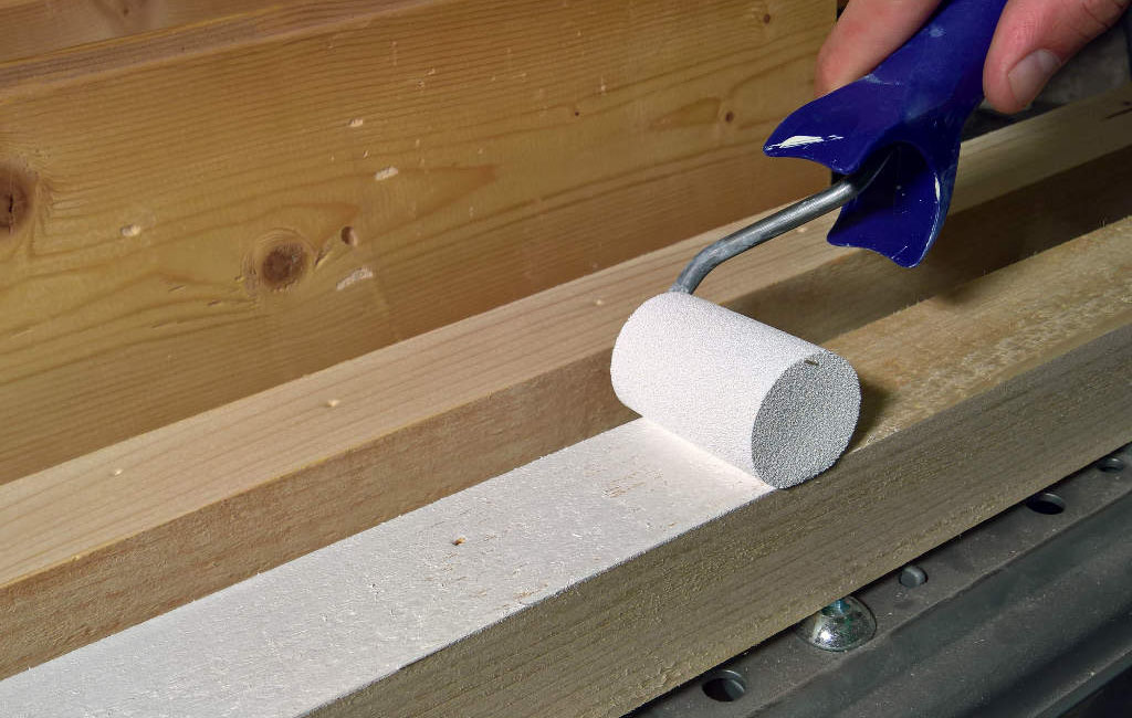 Eine Hand streicht mit einer kleinen Lackrolle weißen Lack auf eine Holzlatte.