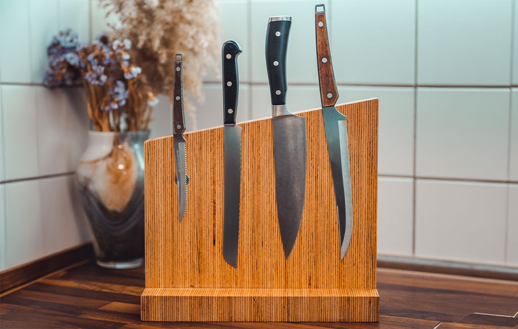 Ein Messerblock aus Holz mit vier verschiedenen Küchenmessern steht auf einer Küchenarbeitsplatte vor weiß gefliester Wand.