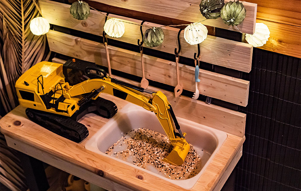 Ein gelber Spielzeugbagger steht auf der Holzplatte der Matschküche und gräbt in einem mit Sand gefüllten Spülbecken.