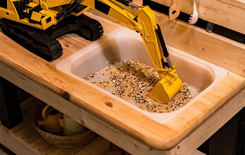 Nahaufnahme der Spielspüle in der Matschküche mit Sand, Körnern und gelbem Spielbagger.