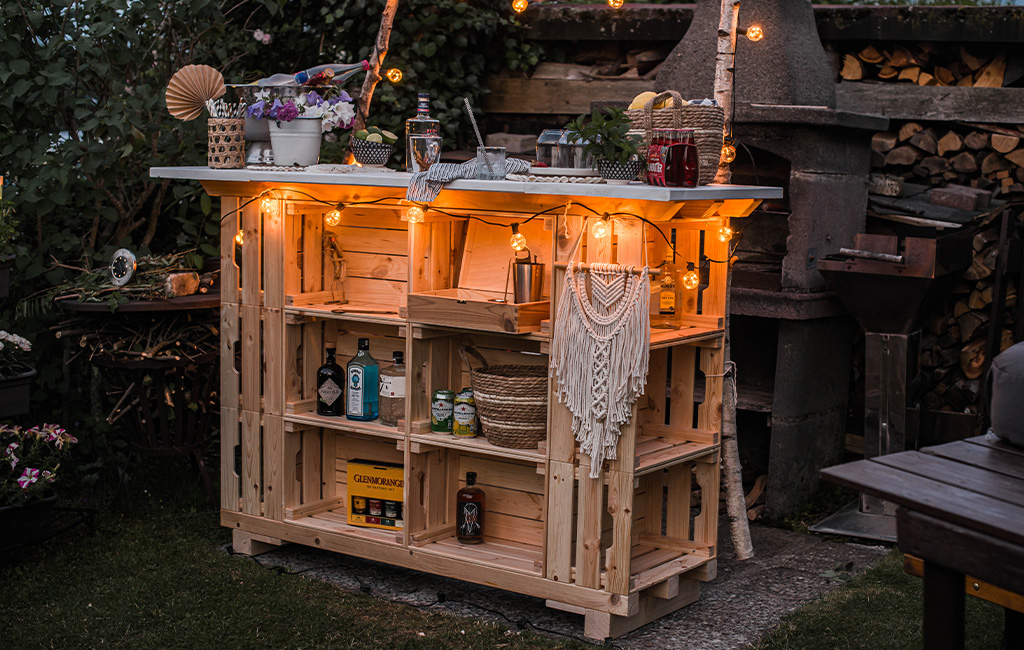 Selbstgebaute Gartenbar aus Holzkisten, dekoriert mit Lichterkette, Pflanzen und Getränken im abendlichen Garten.