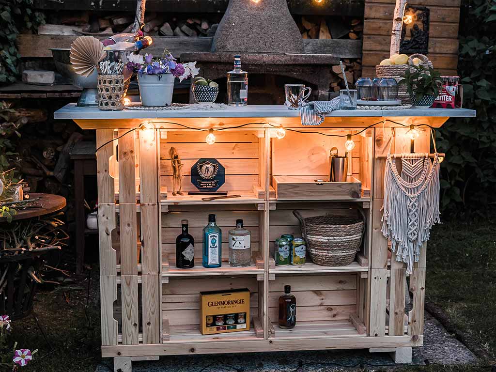 Selbstgebaute Gartenbar aus Holzkisten, dekoriert mit Lichterkette, Pflanzen und Getränken im abendlichen Garten.