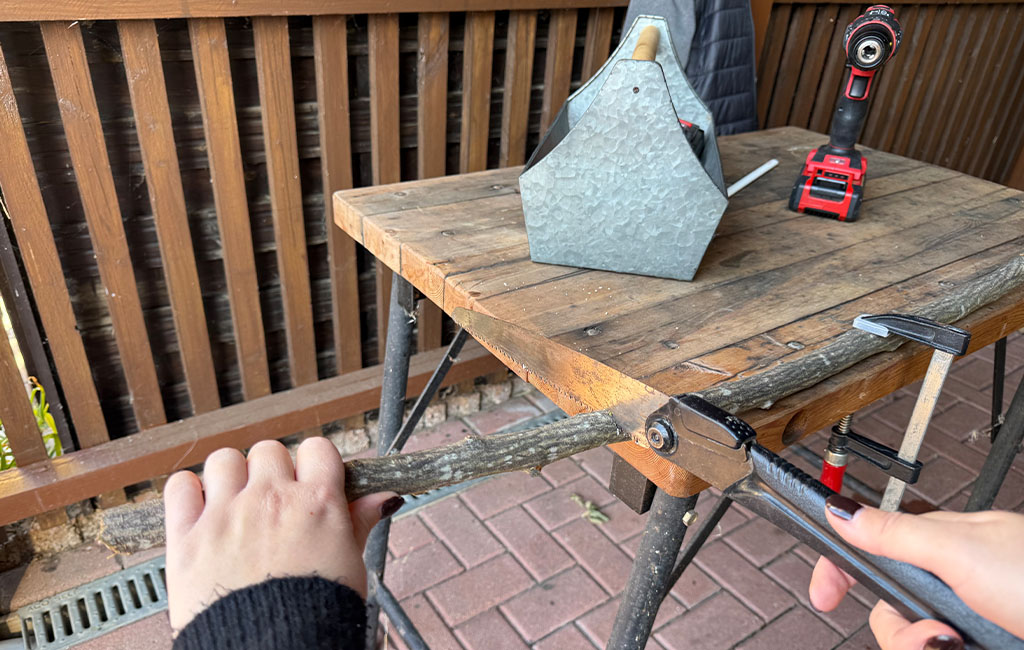 Eine Person sägt einen Ast auf einem Tisch mit einer Handsäge ab, der Ast ist mit einer Zwinge fixiert.