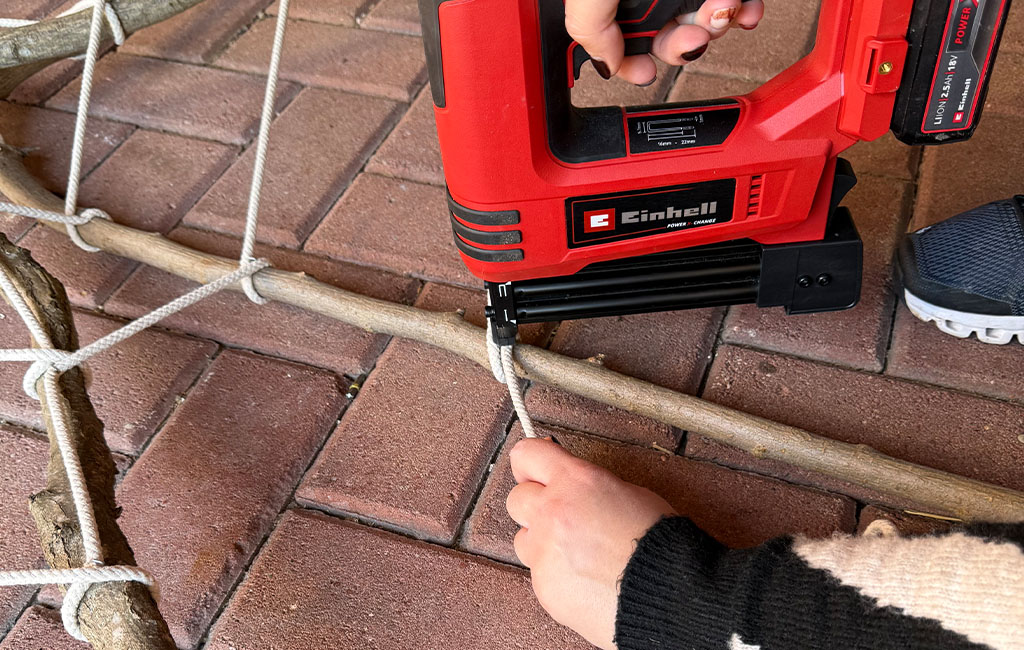 Eine Person befestigt ein Seil mit einem Einhell Akku-Tacker an einem Ast auf dem Boden.