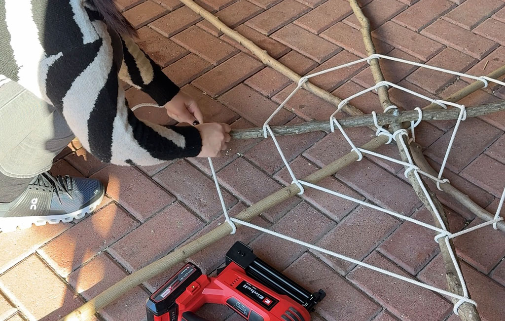 Eine Person knüpft ein Spinnennetz aus Seil an einem Gestell aus Ästen auf dem Boden.