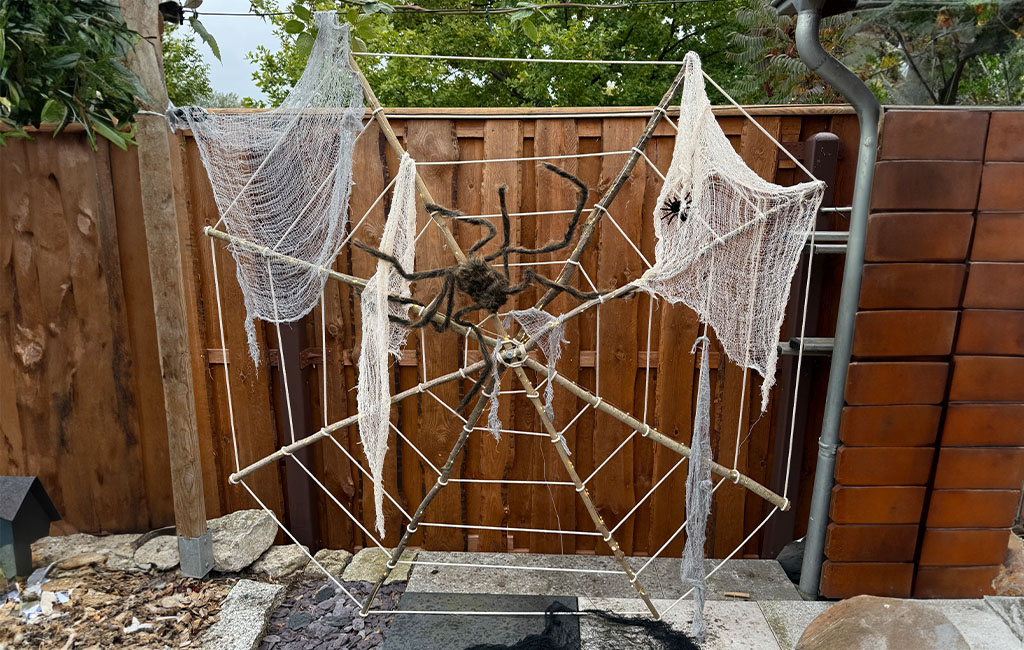Großes Spinnennetz aus Seilen mit weißen Tüchern und zwei künstlichen Spinnen, auf Holzrahmen im Garten montiert.