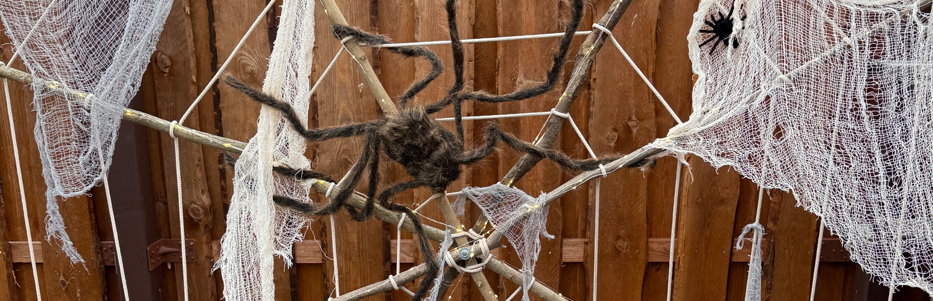 Großes Spinnennetz aus Seilen mit weißen Tüchern und zwei künstlichen Spinnen, auf Holzrahmen im Garten montiert.