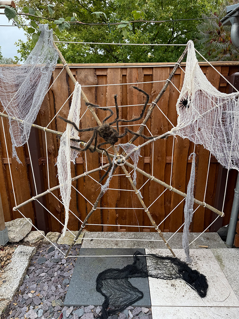 Großes Spinnennetz aus Seilen mit weißen Tüchern und zwei künstlichen Spinnen, auf Holzrahmen im Garten montiert.